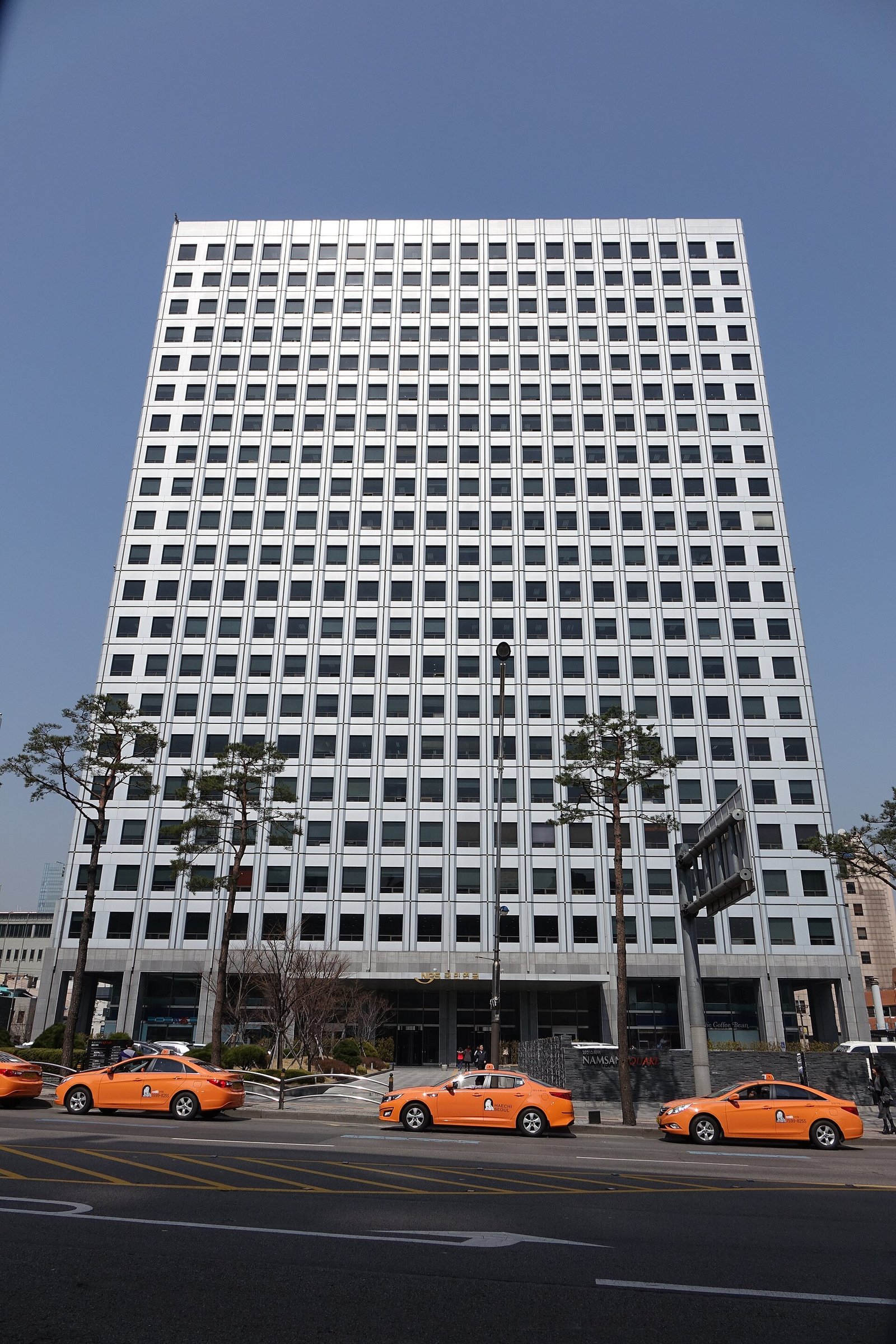 맑은 하늘 아래 창문이 촘촘한 흰색 고층 건물이 서 있고, 앞 도로에 주황색 택시 여러 대가 서 있다. 국민연금 건물이다