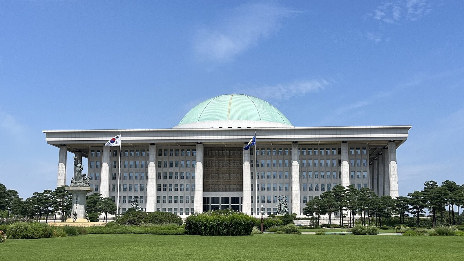 맑은 여름 하늘 아래 초록색 돔 지붕과 흰 기둥이 있는 국회의사당 건물. 앞에 잔디밭과 소나무가 펼쳐져 있다