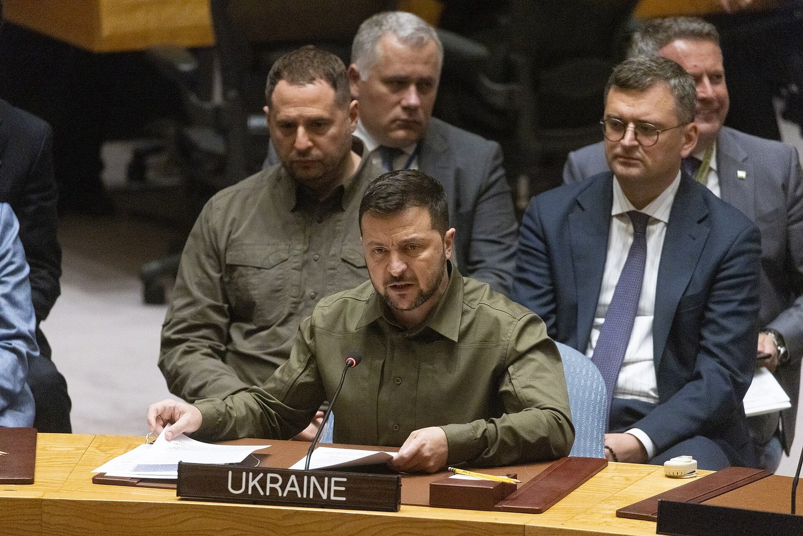 올리브색 셔츠를 입은 젤렌스키 우크라이나 대통령이 UKRAINE 명패가 놓인 유엔 안보리 회의석에서 마이크 앞에 앉아 연설하고, 뒤로 수행원들이 앉아 있다
