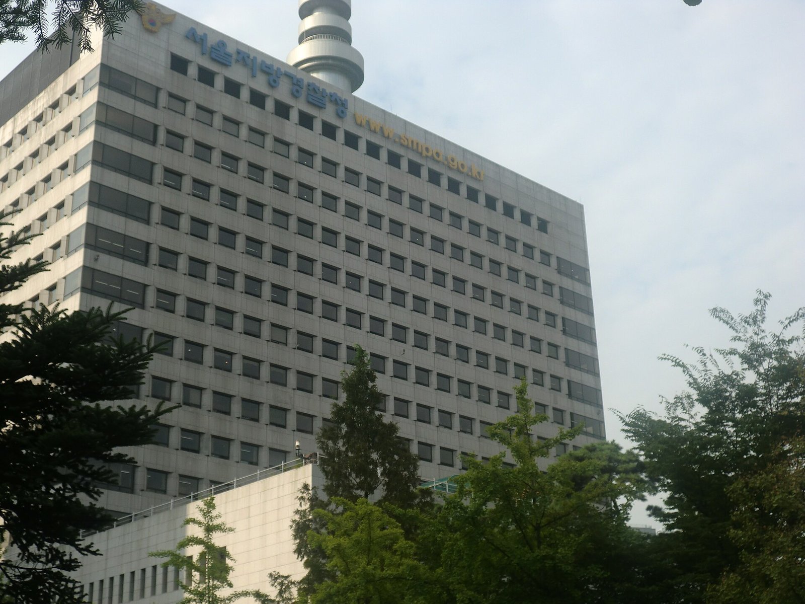 나무 사이로 올려다본 회색 고층 건물 위쪽에 파란 글씨로 '서울지방경찰청'과 노란 글씨로 'www.smpa.go.kr'이 적혀 있고, 뒤로 원통형 탑이 보인다