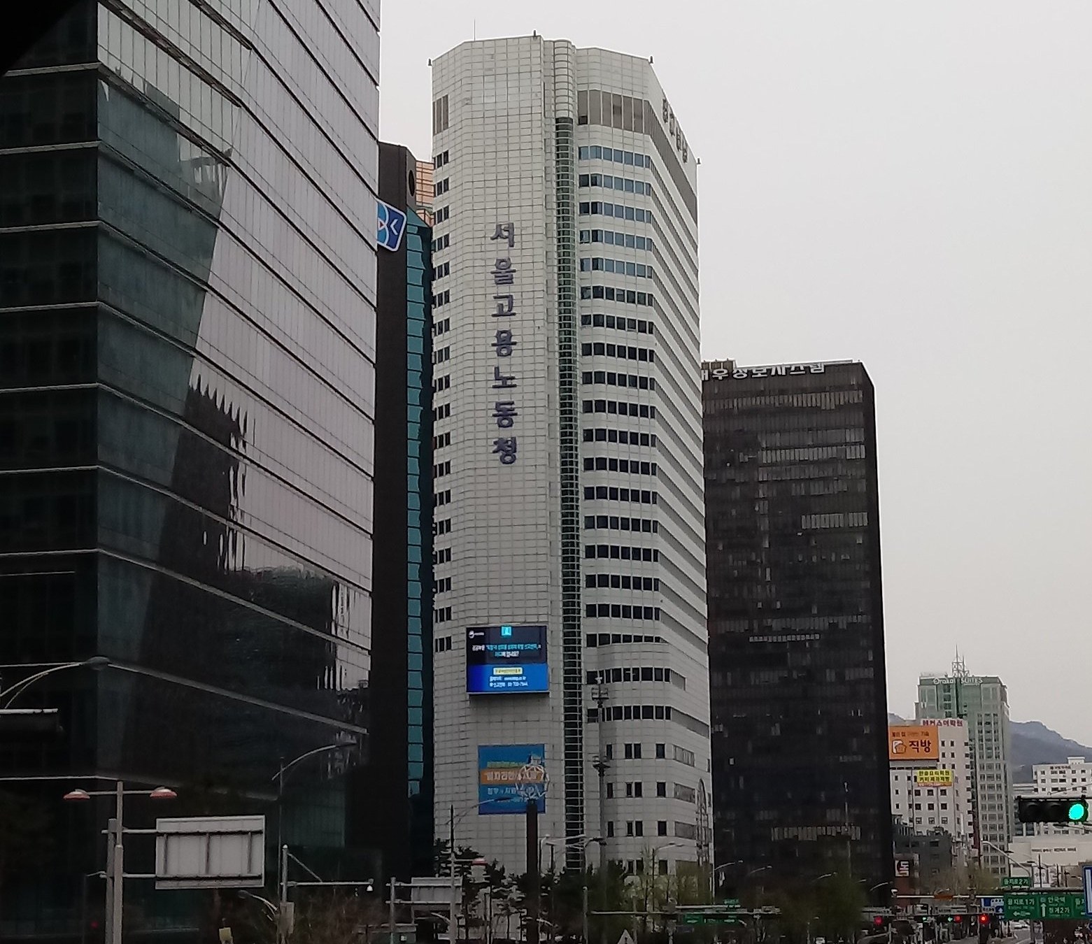흐린 하늘 아래 고층 빌딩들 사이로 세로로 '서울고용노동청'이라고 적힌 흰색 건물이 서 있다