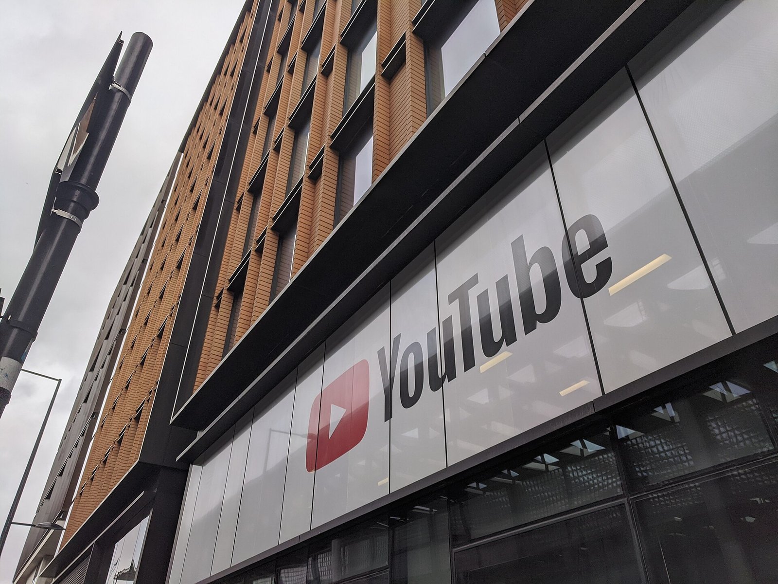 흐린 하늘 아래 올려다본 건물 외벽에 빨간 재생 버튼과 YouTube 로고가 크게 붙어 있다. 위층은 주황색 벽돌 기둥이 줄지어 있다